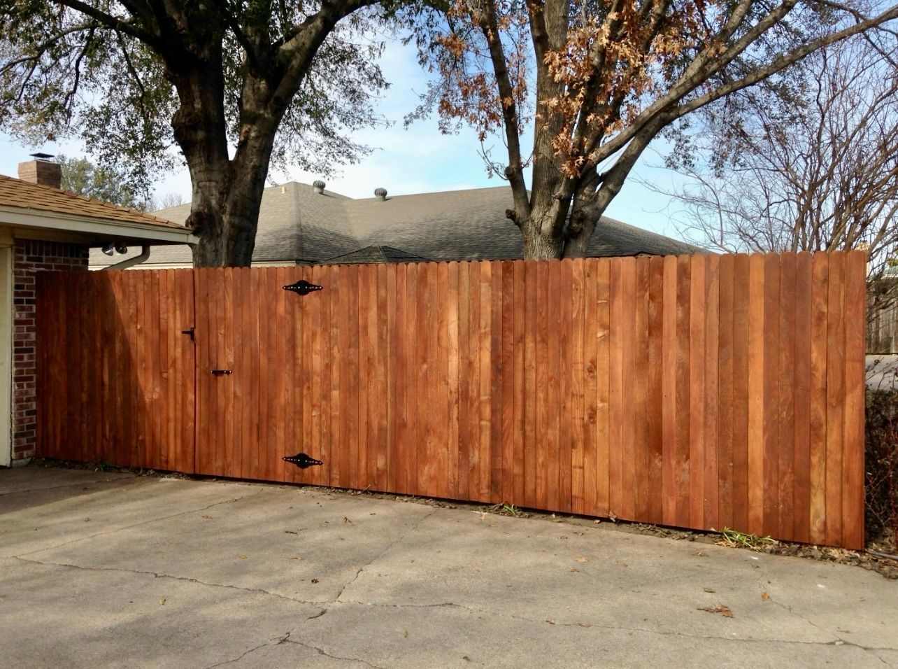 Brown wooden fence with black hinges, near a brick wall and trees.