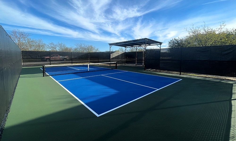 Blue pickleball court with black fencing, blue sky, and a covered seating area.
