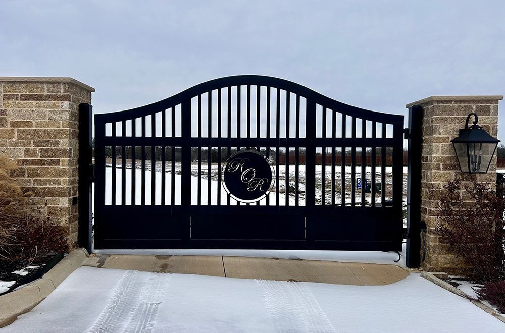 Black metal driveway gate with decorative top and monogram, flanked by brick columns and snow.