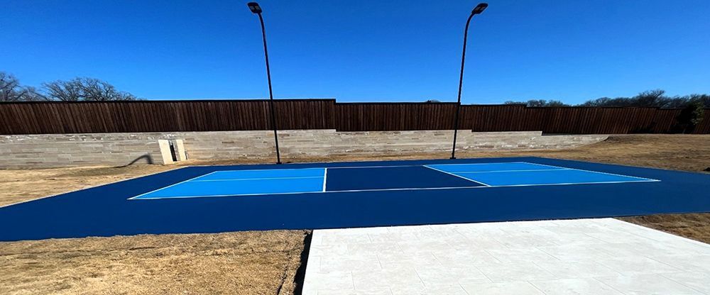 Blue pickleball court with white lines and lights, set against a fence and clear blue sky.