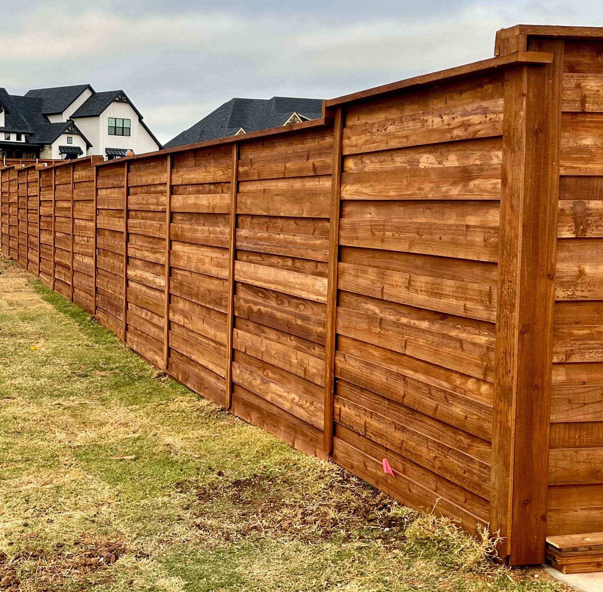 Wooden privacy fence stained brown, extending along a grassy yard with houses in the background.