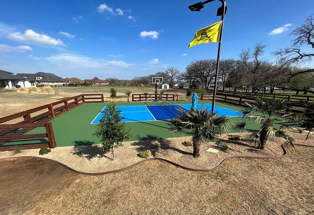 Basketball court with blue and green surface, fenced and landscaped, with a flag waving in the sky.
