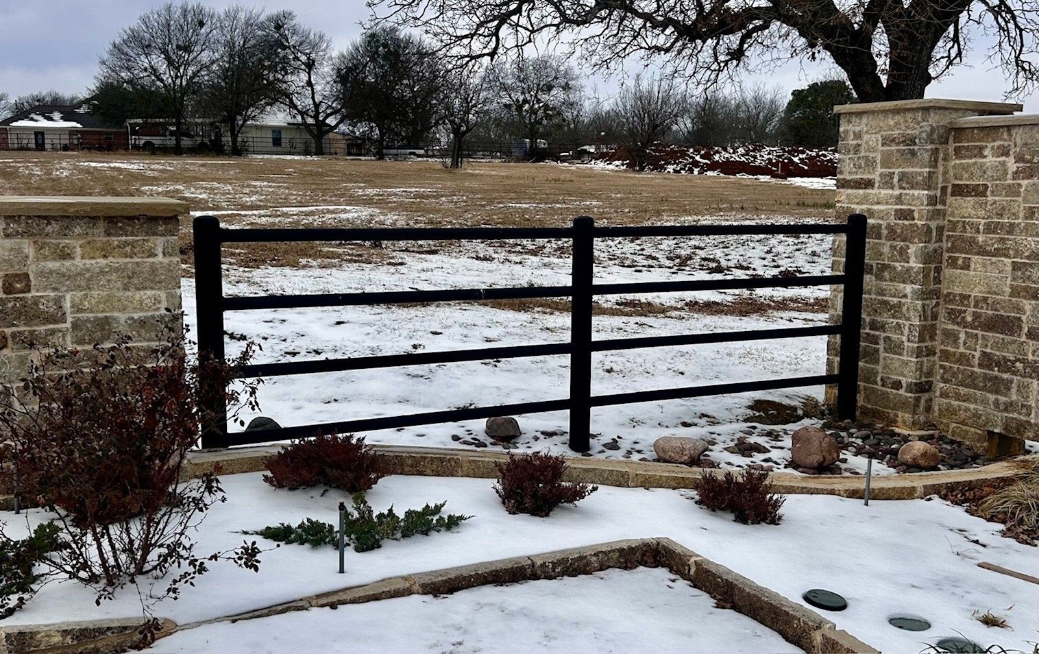 Black metal fence with stone pillars and snow-covered ground in a rural setting.