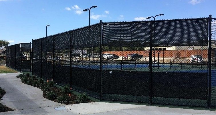 Black chain-link fence surrounds a blue tennis court, with a paved walkway and landscaping in the foreground.