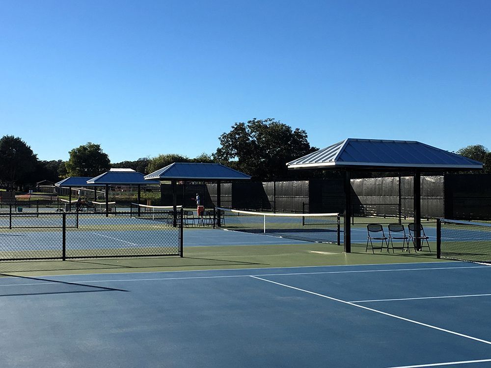 Outdoor tennis courts with blue surfaces, nets, and shaded benches under a clear blue sky.