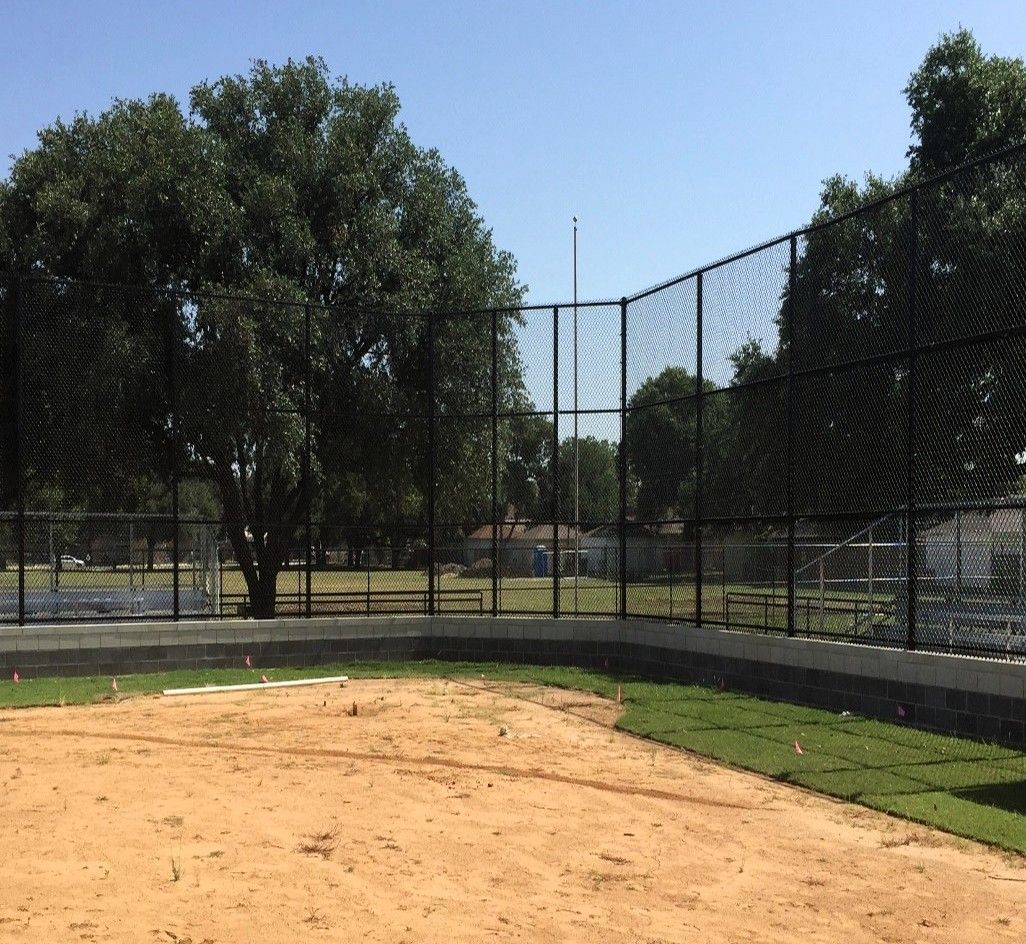 Baseball field with a chain-link backstop, dirt infield, and green grass. Trees in the background.
