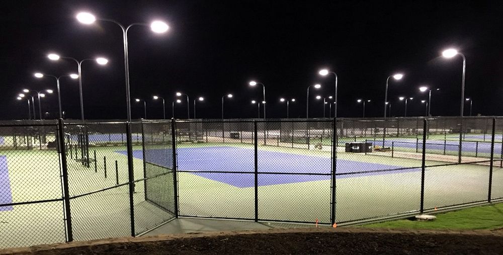 Night view of illuminated pickleball courts behind a black fence.