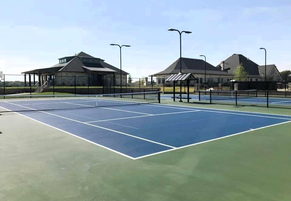 Tennis court with blue playing surface, green surround, and buildings in the background.
