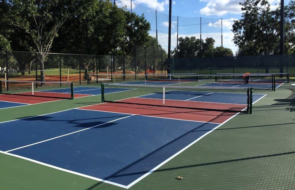 Pickleball courts with blue, red, and green surfaces, net, fence, and trees under a partly cloudy sky.