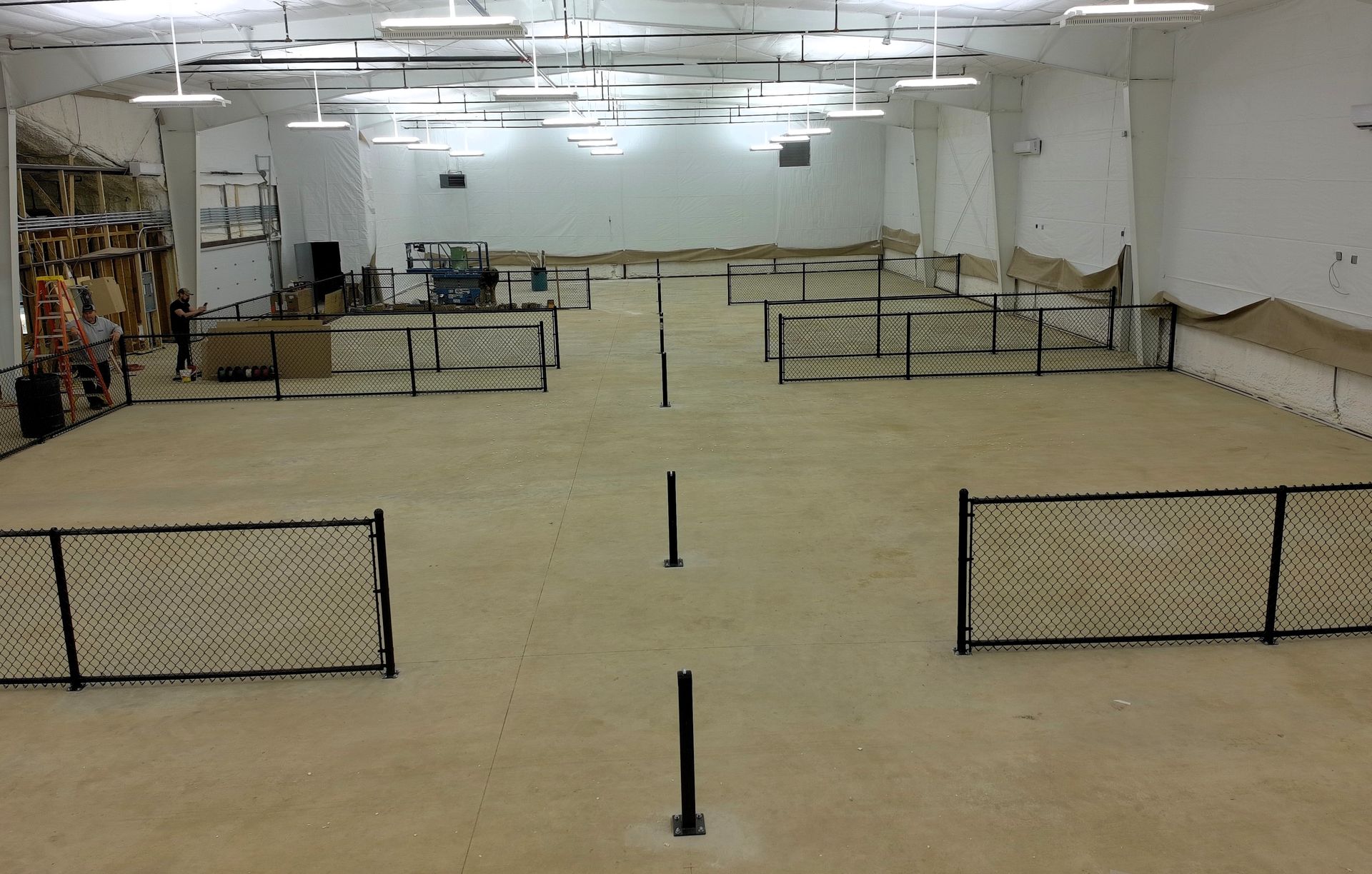 An empty warehouse interior with tan-colored flooring and black metal fences. Overhead lights illuminate the space.