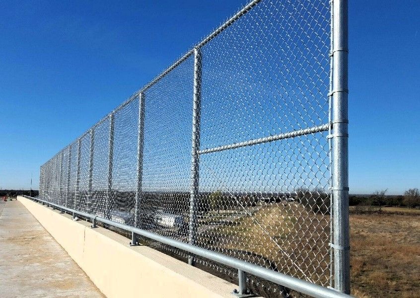 Chain-link fence atop a concrete barrier, overlooking a highway and landscape under a blue sky.