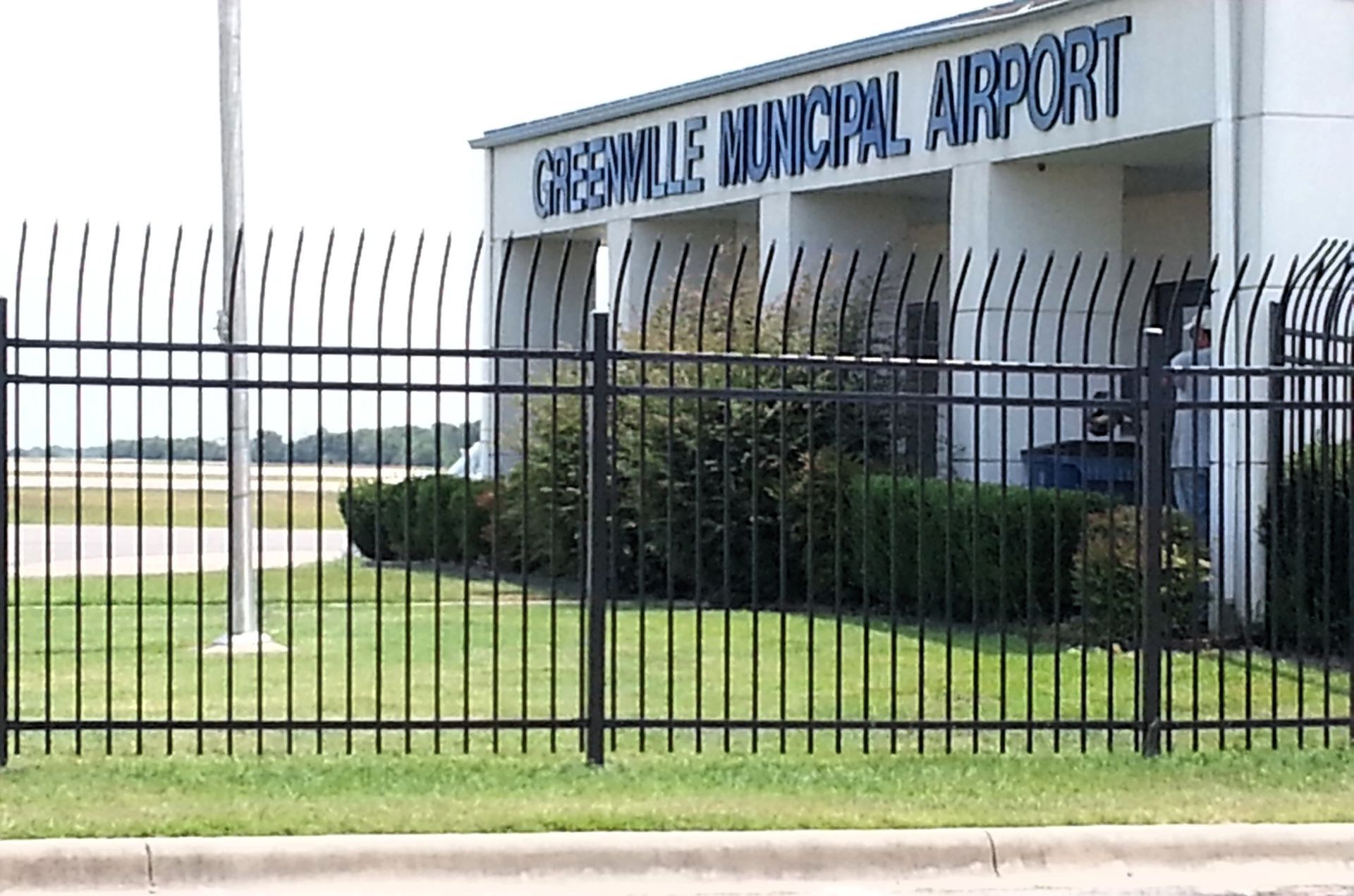 Greenville Municipal Airport building behind a black metal fence, with green grass and sky visible.