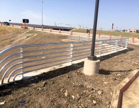 Metal guardrail along a concrete walkway next to a road, dirt and a light pole.