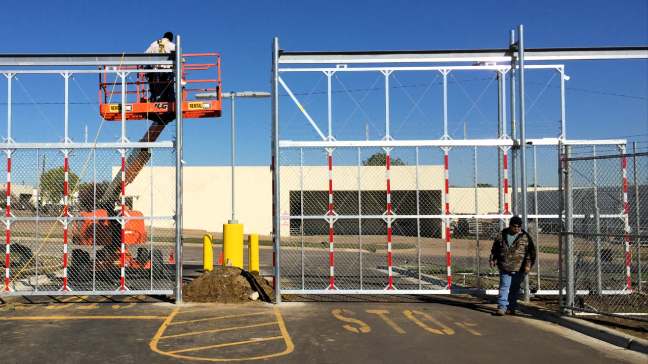Workers install gate on fence with lift in front of a building; one person stands by.