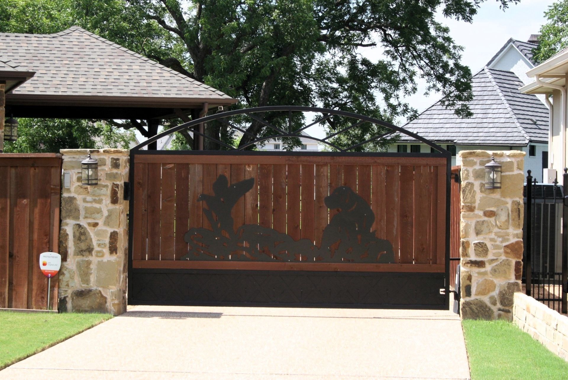 Wooden gate with decorative cutout of a landscape, between stone pillars, over a driveway.