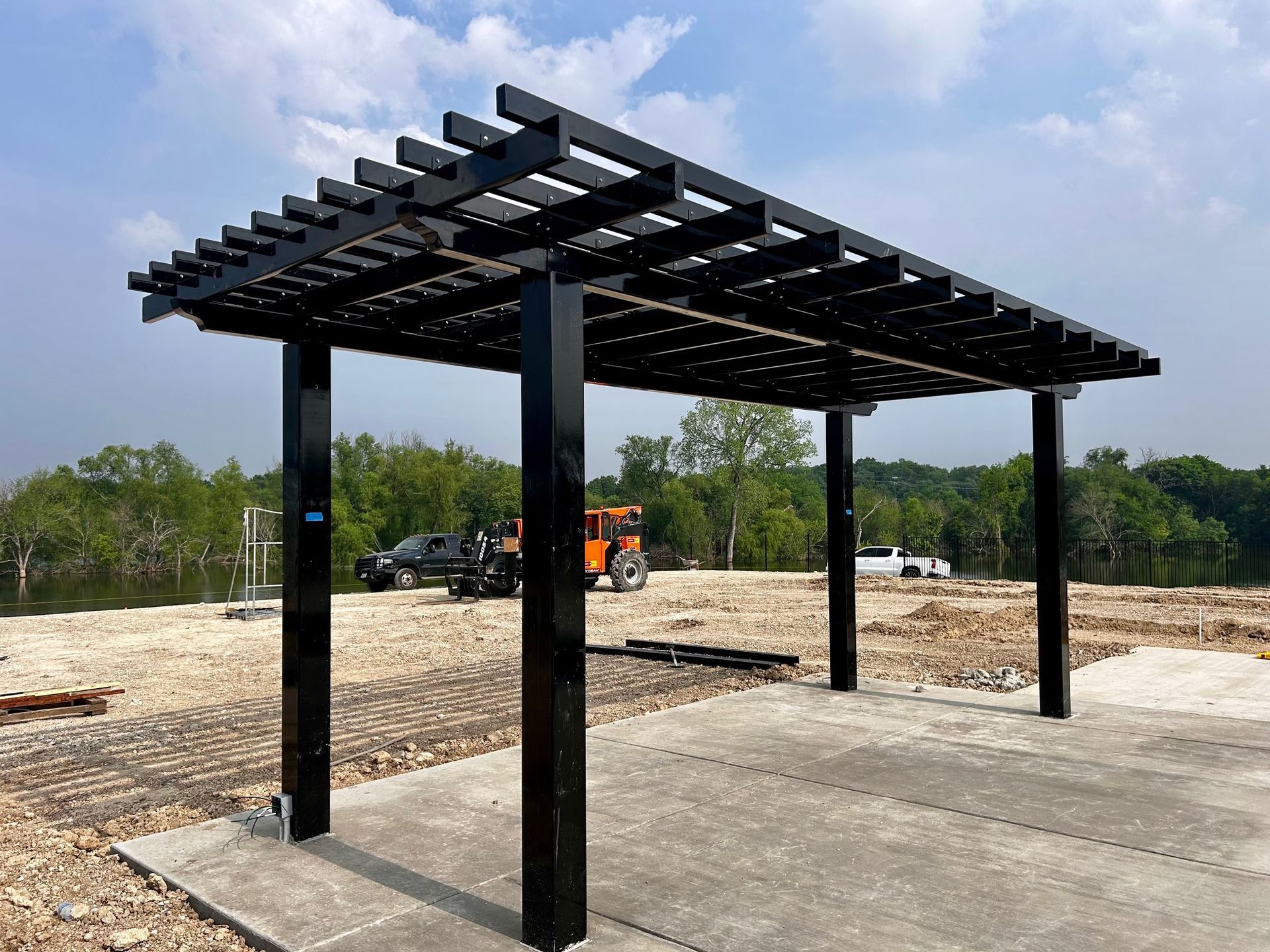 Black pergola with horizontal slats over a concrete pad, on a partially cleared lot with trees in the background.