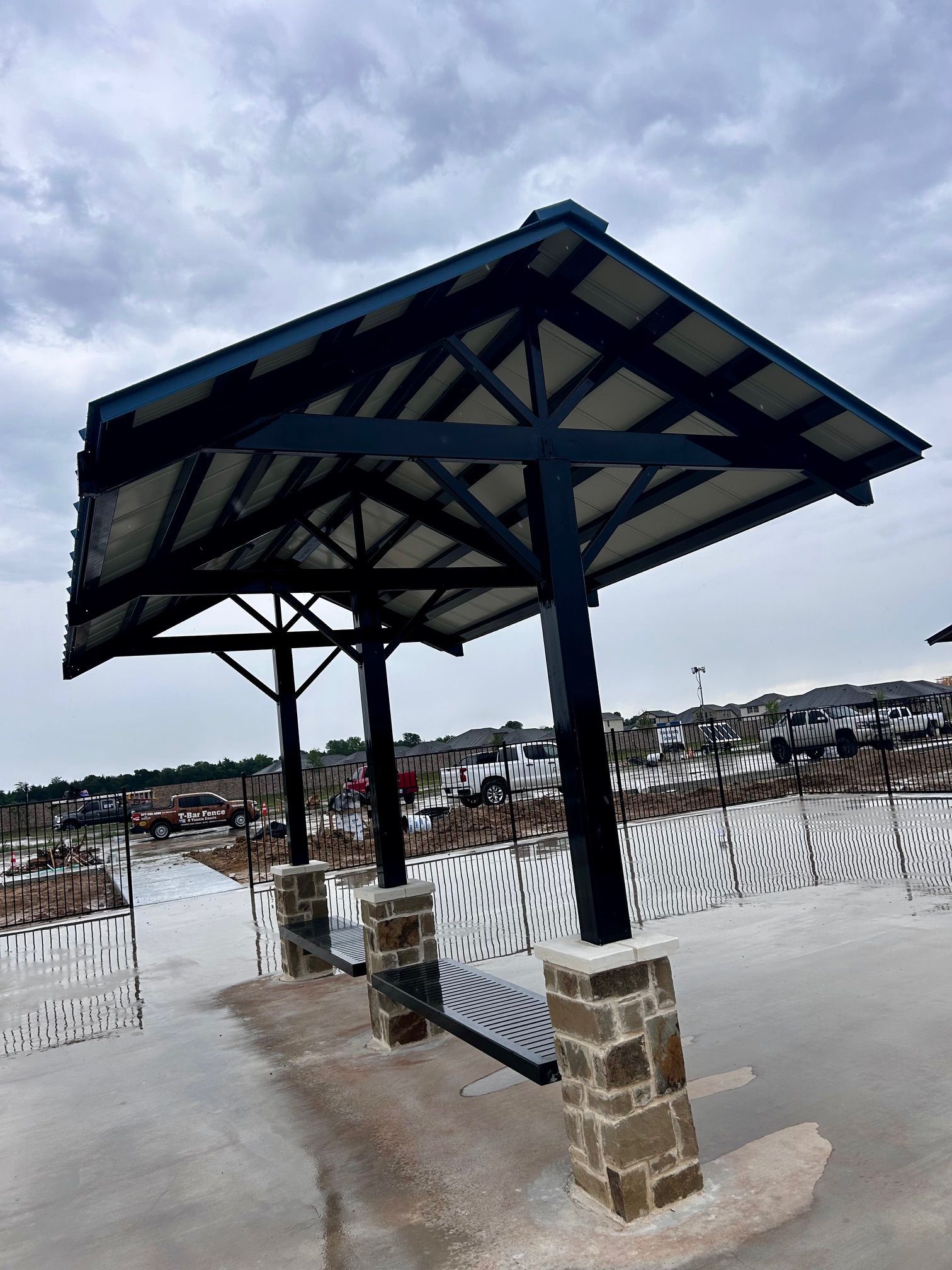 Park shelter with black frame, gray roof, stone pillars, and bench on a concrete platform against a cloudy sky.