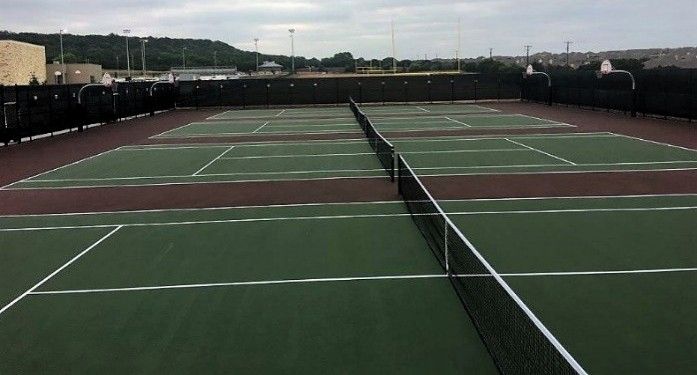 Several green pickleball courts with black nets, brown surrounds, and a background of trees under a cloudy sky.