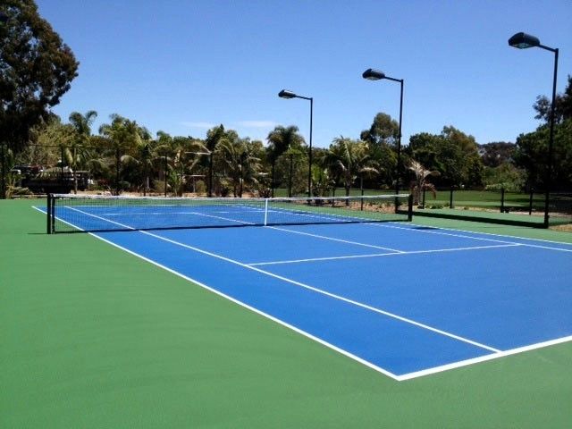 Blue and green tennis court with net, lights, and trees under a blue sky.
