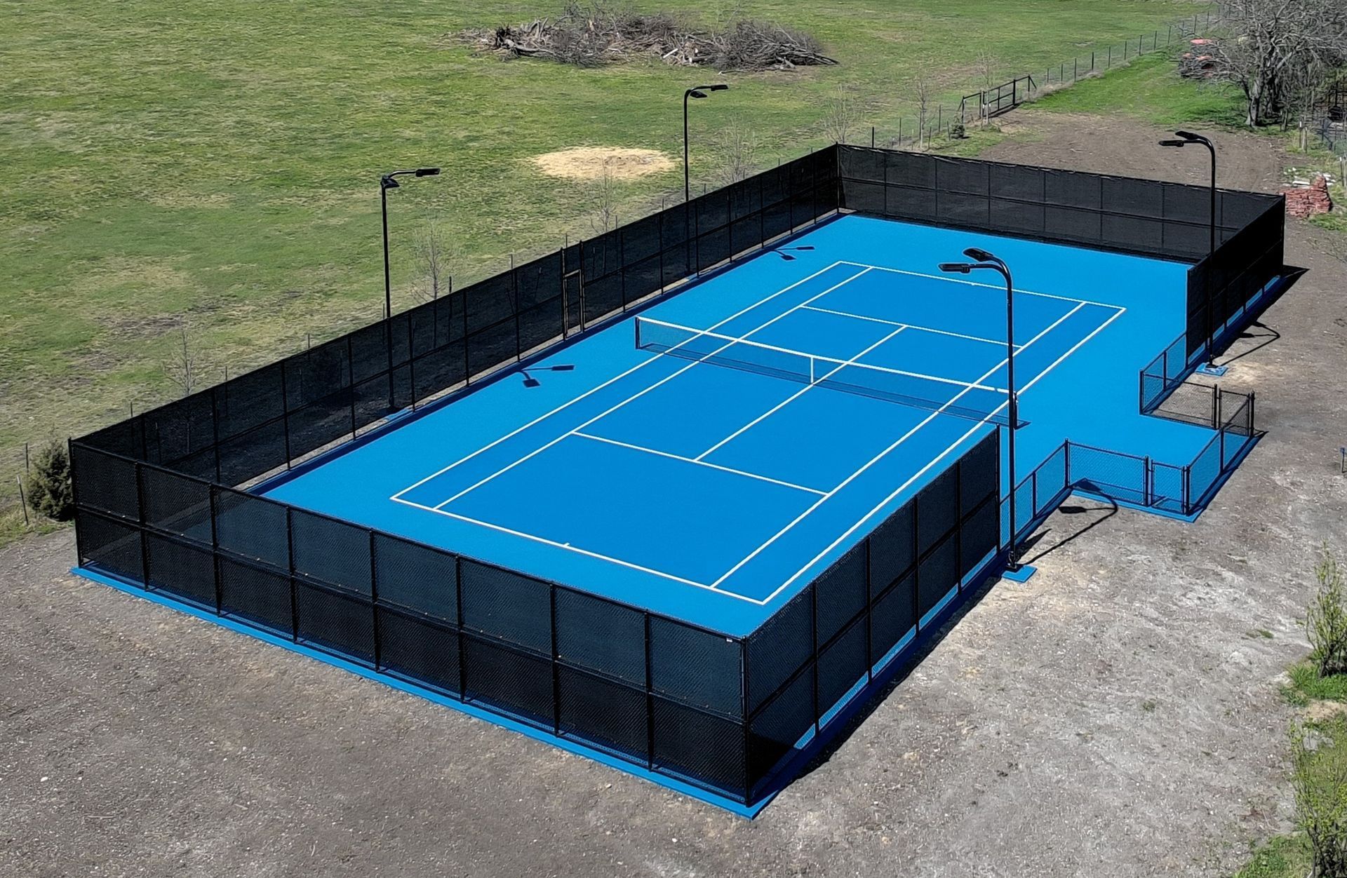 A bright blue outdoor tennis court surrounded by a tall black chain-link fence, set in a grassy, undeveloped area.