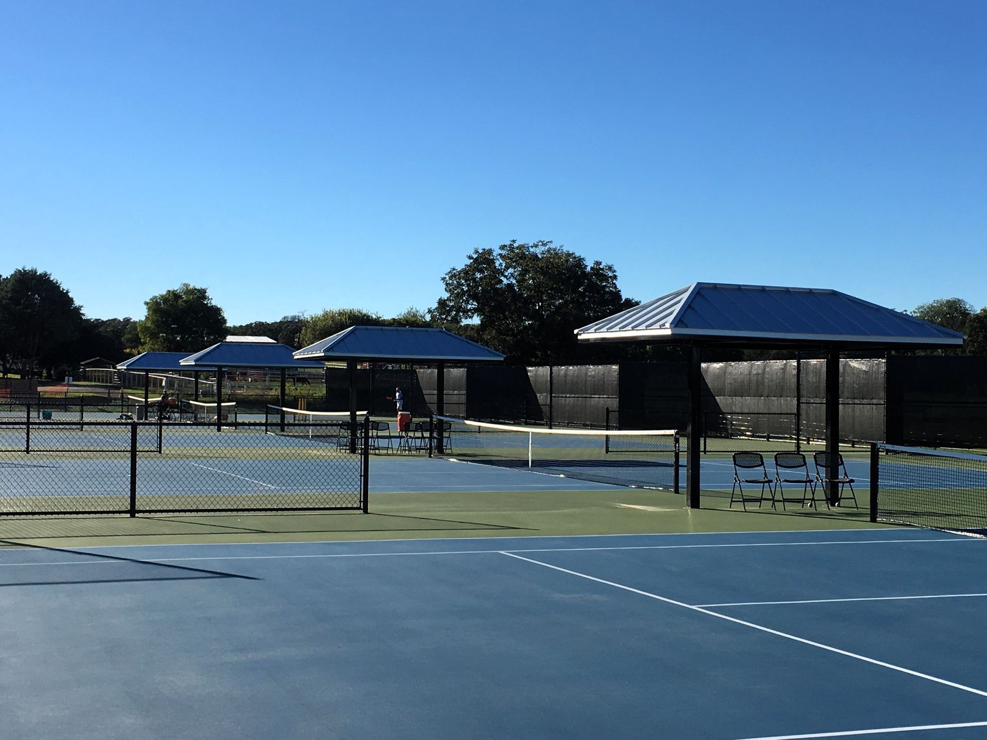 A sunny outdoor blue tennis court with several shaded bench areas along the fence line.