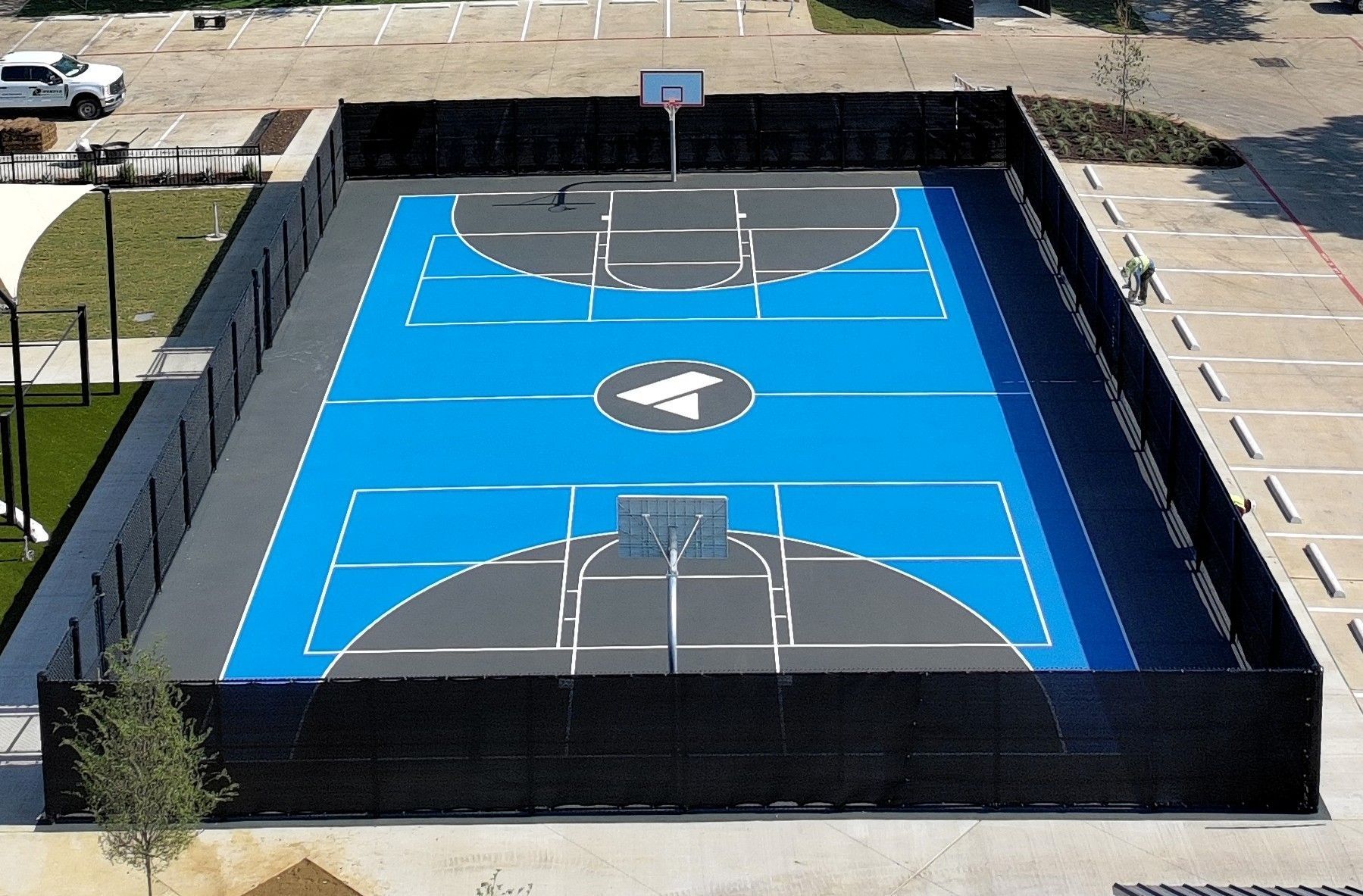 Aerial view of a blue outdoor basketball court with black fencing and white lines.