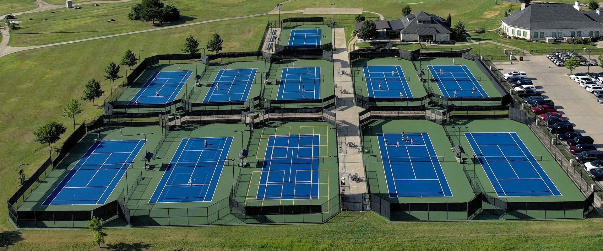 Aerial view of outdoor blue tennis courts in a fenced complex beside grassy fields