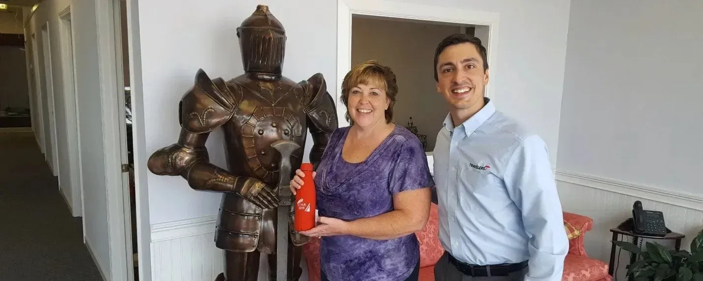 A woman and a man stand in a room next to a suit of armor; the woman holds a small red object.
