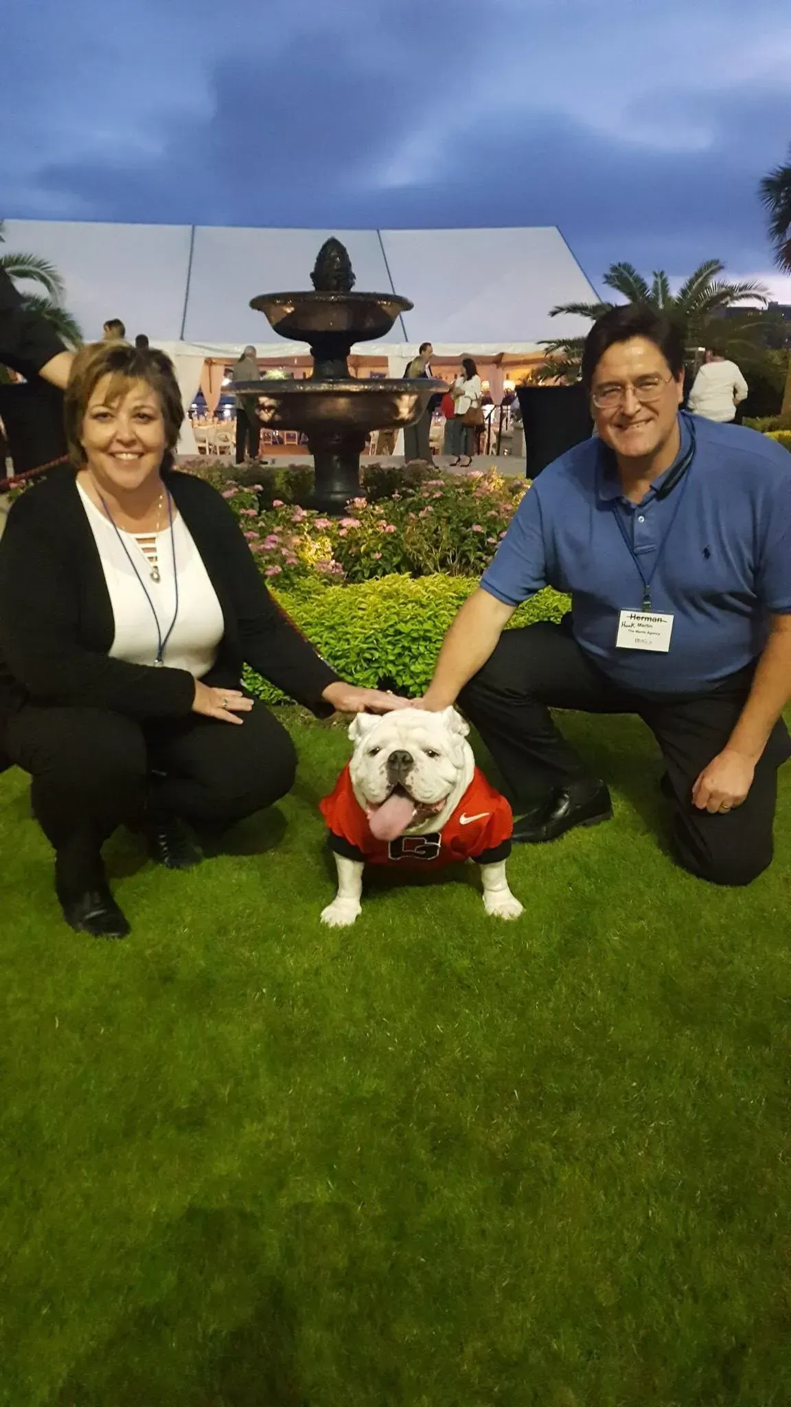 A woman and a man kneel on grass beside a bulldog wearing a red shirt, with a fountain and tent in the background.