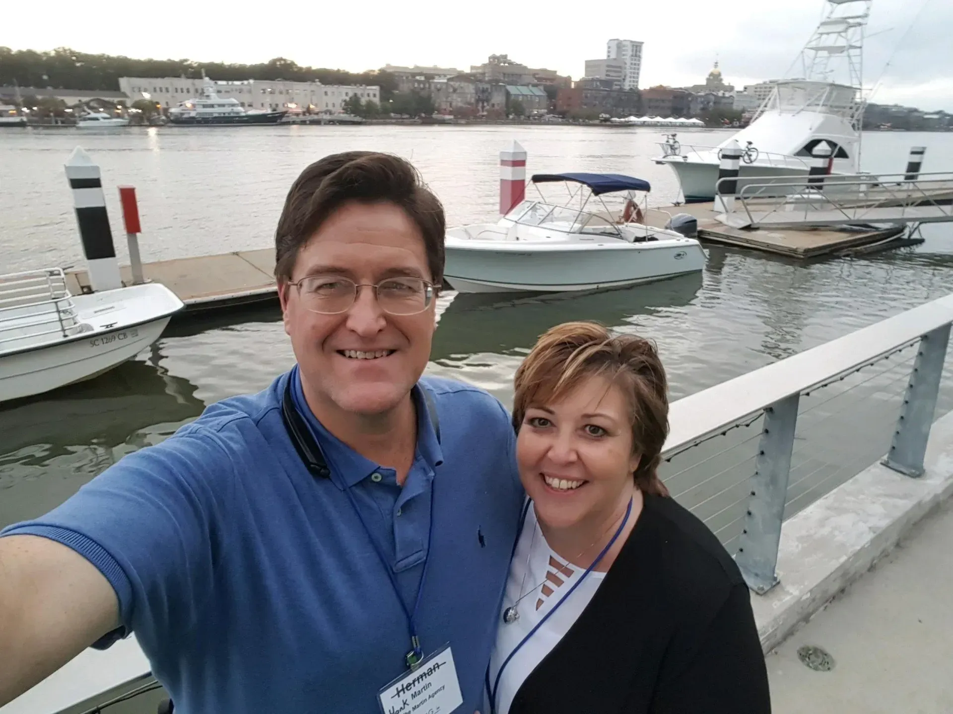 A smiling couple takes a selfie on a waterfront dock with boats and city buildings in the background.
