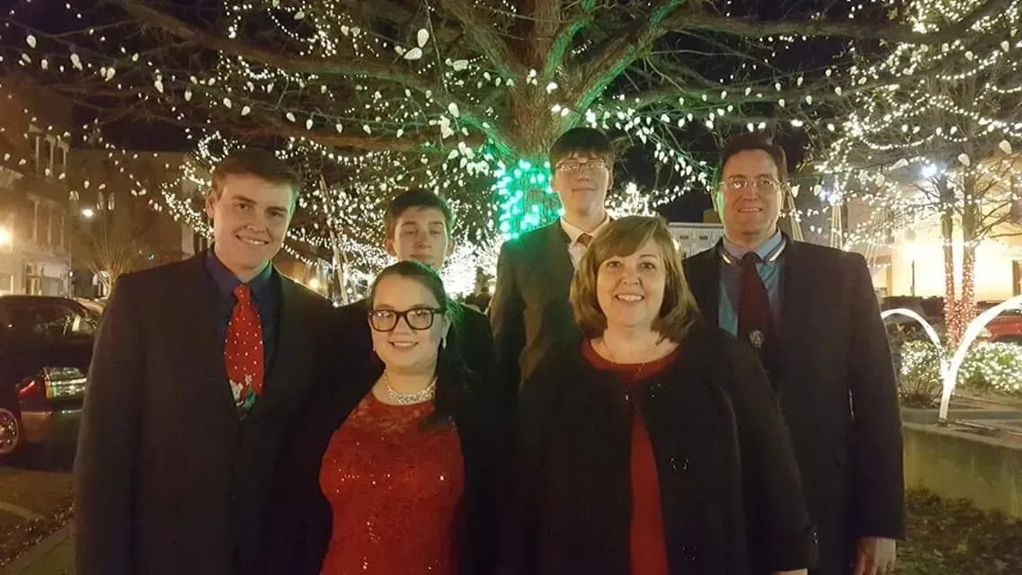 Six people in formal attire pose for a photo at night in front of a tree decorated with bright holiday lights.