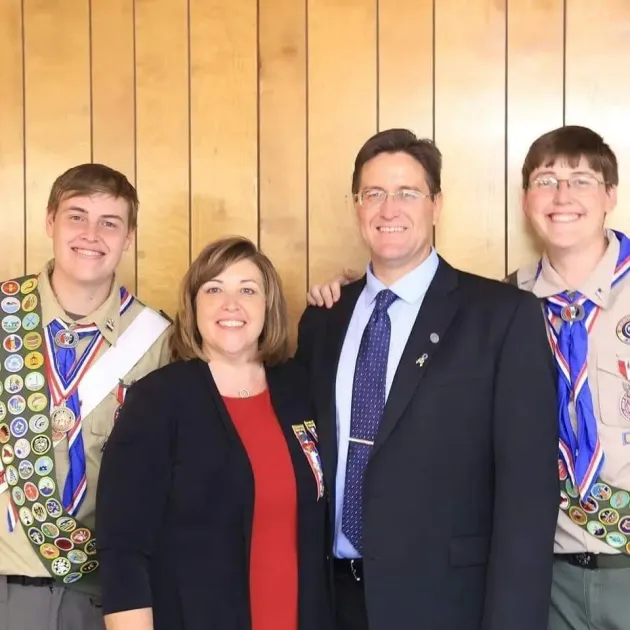 A woman and man stand between two youth in scouting uniforms with merit badge sashes against a wood-paneled wall.