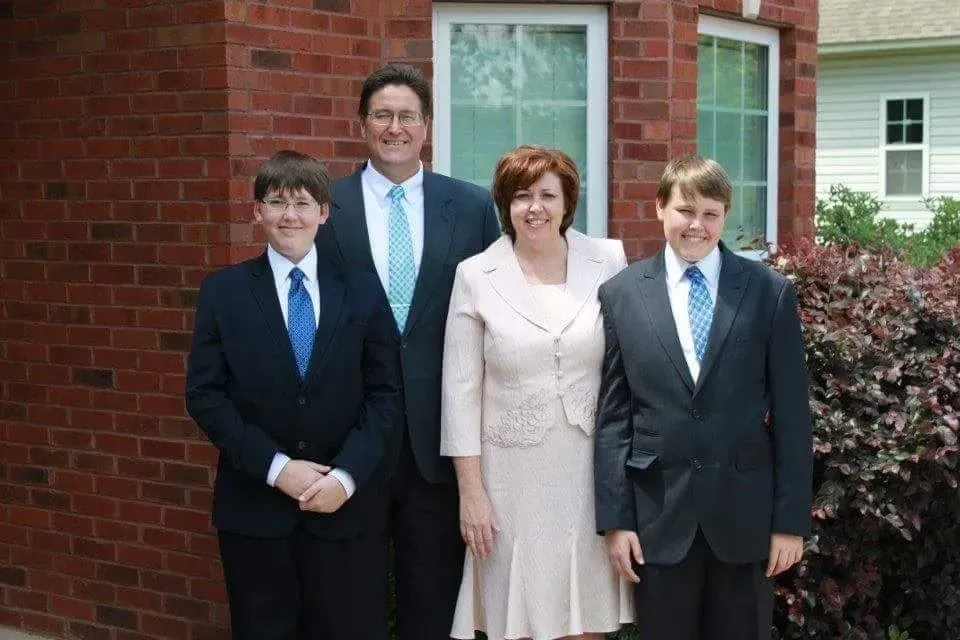 A family of four stands outdoors in front of a brick house, all smiling while wearing formal attire.
