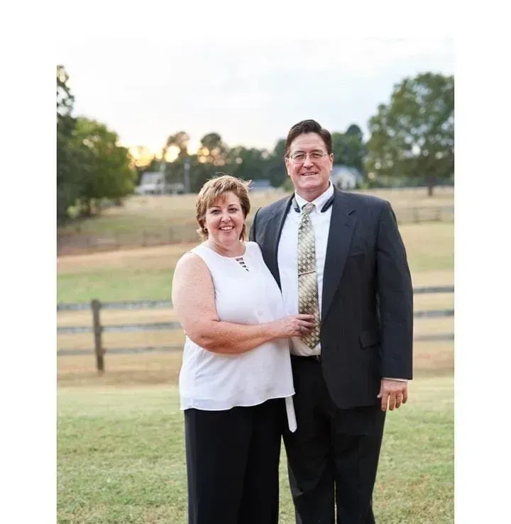 A smiling couple standing outdoors in front of a field and a wooden fence during the golden hour.