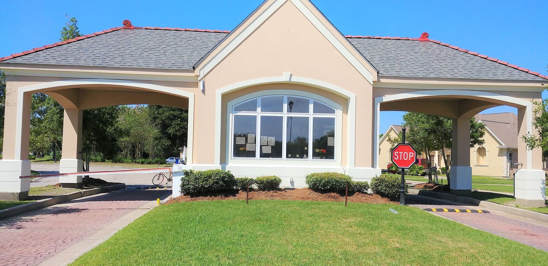 A tan and gray gatehouse with arched entryways, a large window, and a stop sign on a green lawn under a clear blue sky.