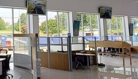Interior of an office with cubicles, windows, and monitors showing car footage. Sunlight streams in.