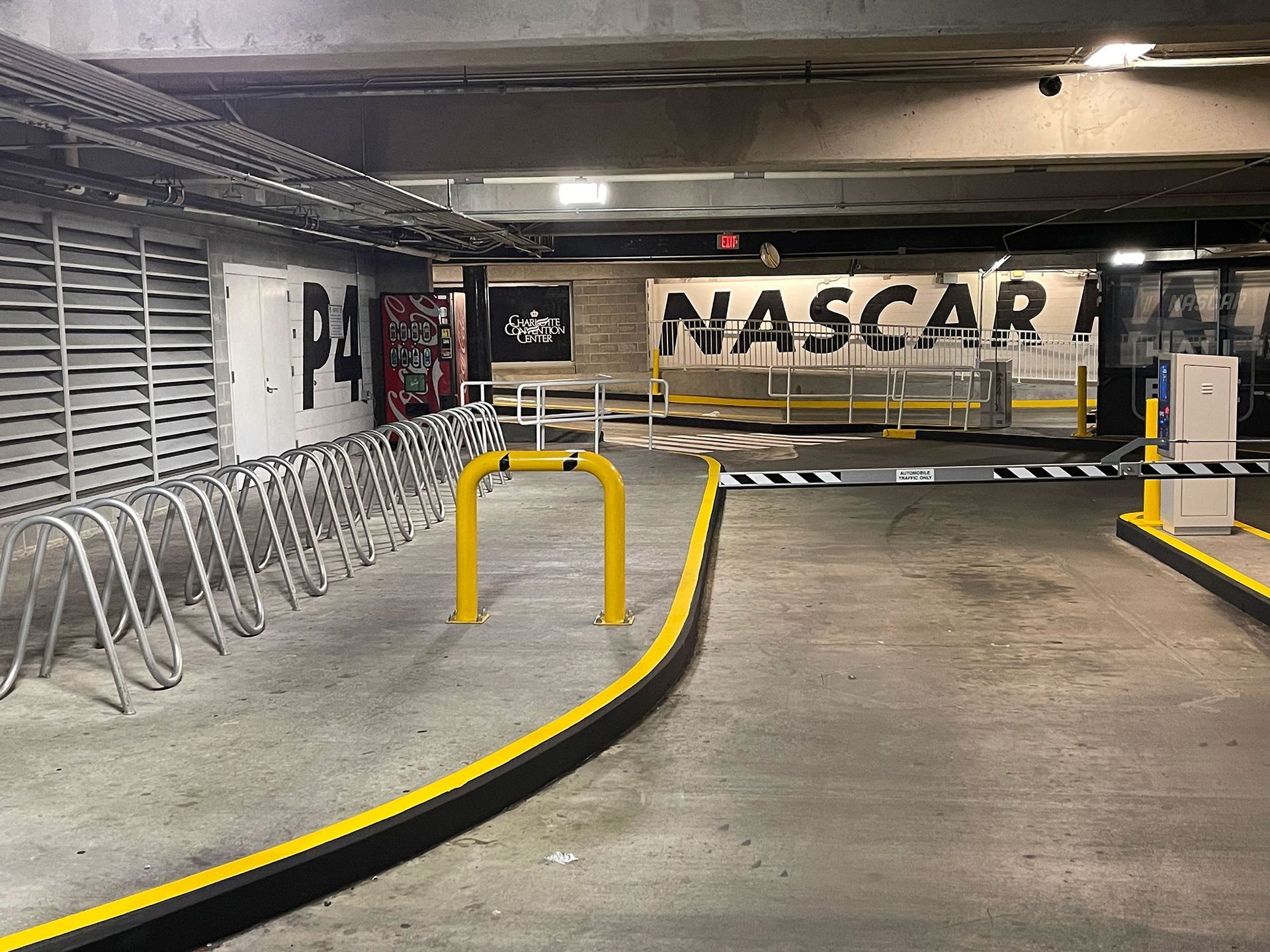 A parking garage entrance with signage for NASCAR and a bicycle rack. The entrance has a yellow safety bar and yellow curbing.