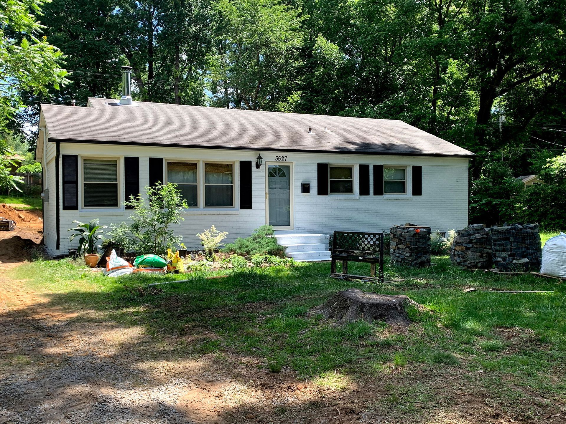 White bungalow house with black shutters, overgrown front yard, and trees in the background.