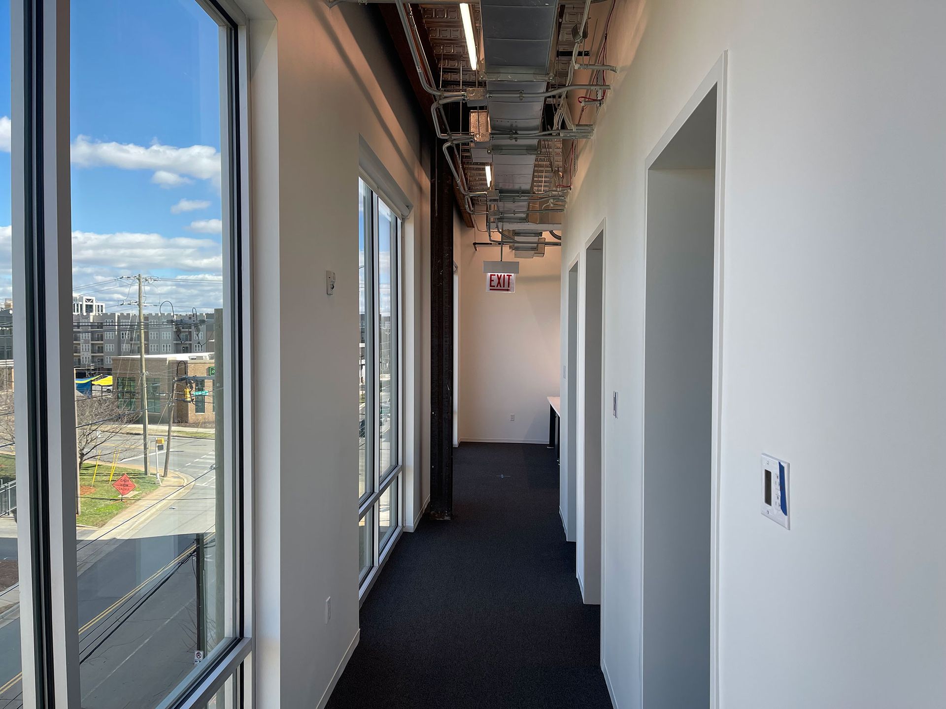Hallway with floor-to-ceiling windows showing a cityscape.  Doors line the right wall, with exposed conduits above.
