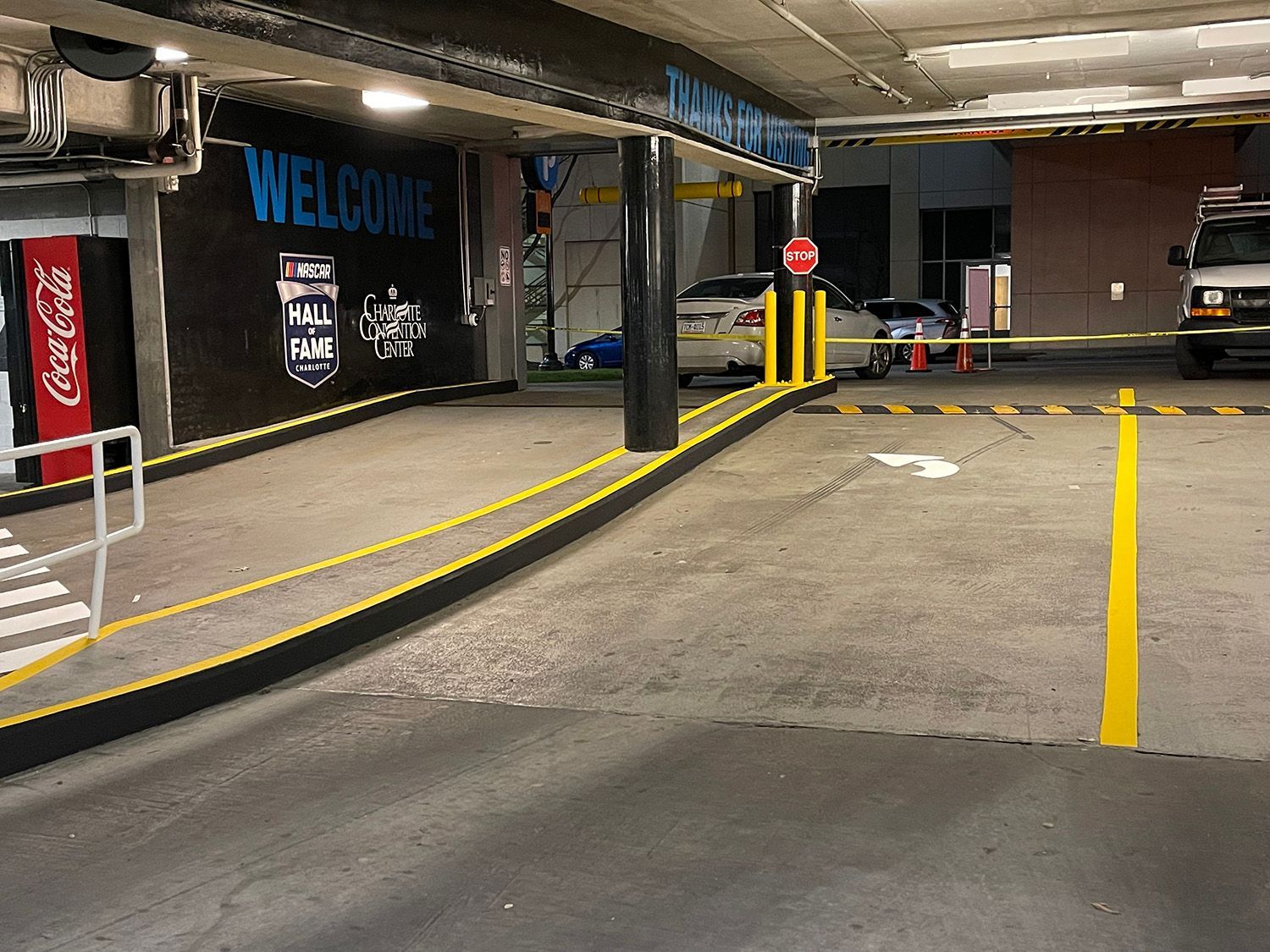 Entrance to an underground parking garage with a Coca-Cola machine, yellow bollards, and parked cars. 