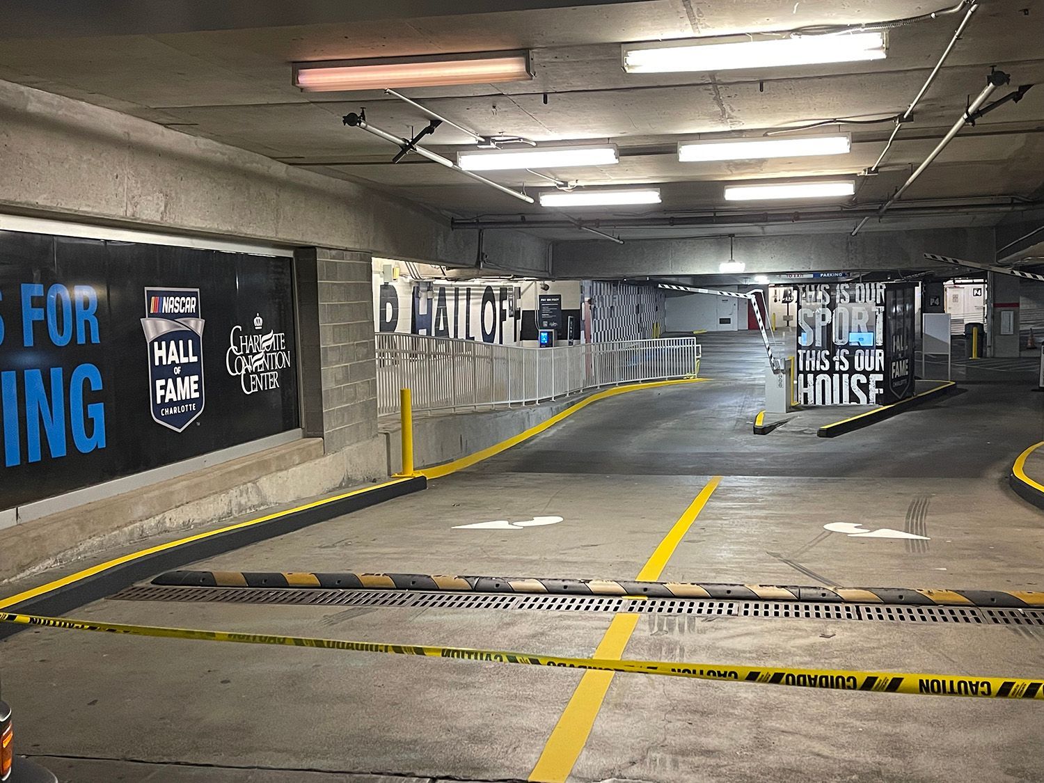 Interior view of a parking garage with a ramp and yellow caution tape. Signs and overhead lights are visible.
