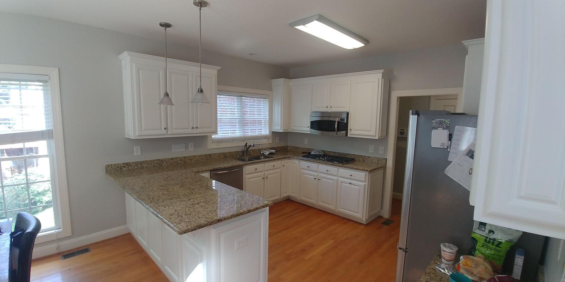 A kitchen with white cabinets, granite countertops, and wooden floors. A stainless steel refrigerator is on the right.