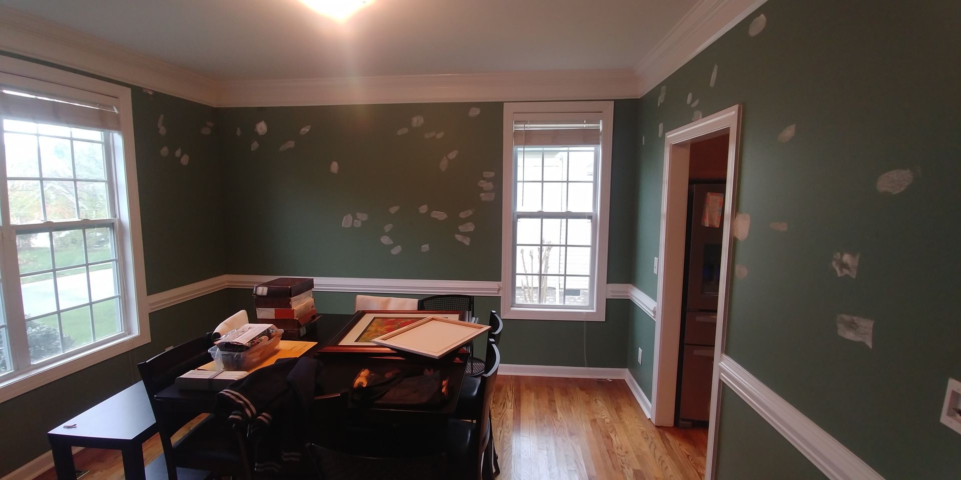 A dining room with dark green walls and white trim, a dark table, and wooden floors. Patches are visible on the walls.