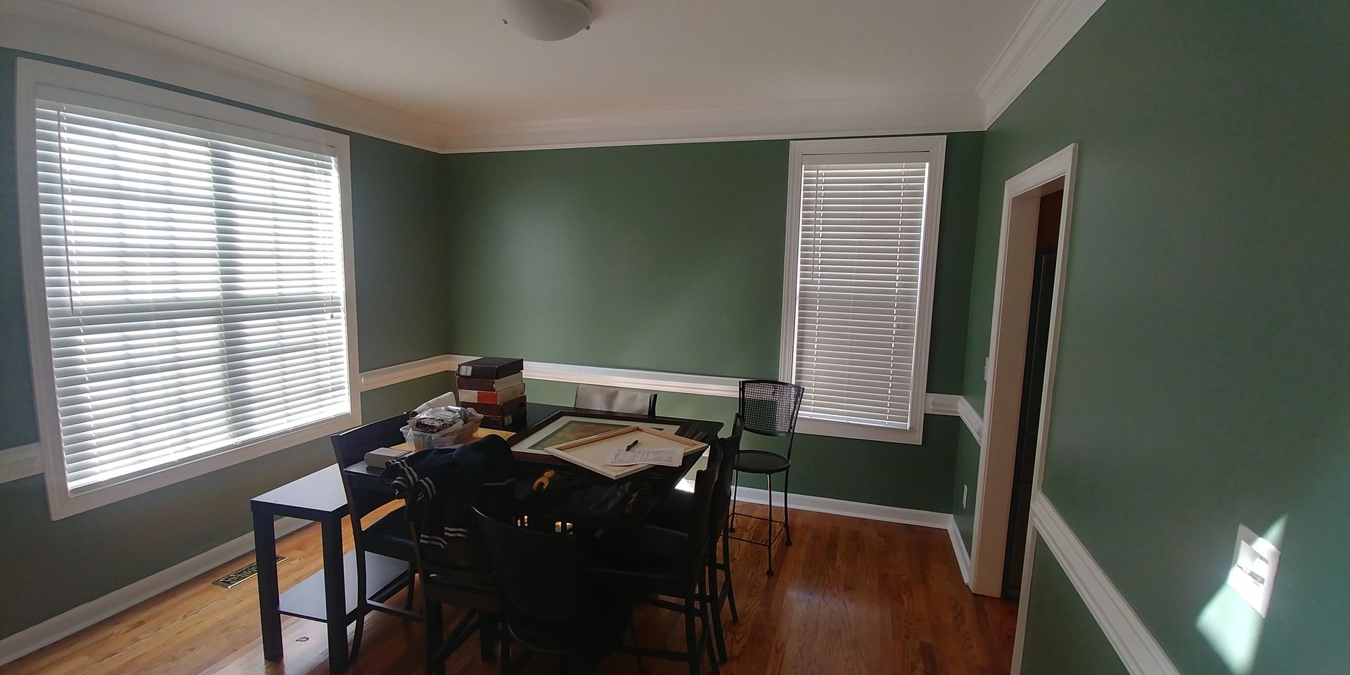 A dining room with green walls, white trim, and a table set for a game. Sunlight streams through the blinds on the windows.