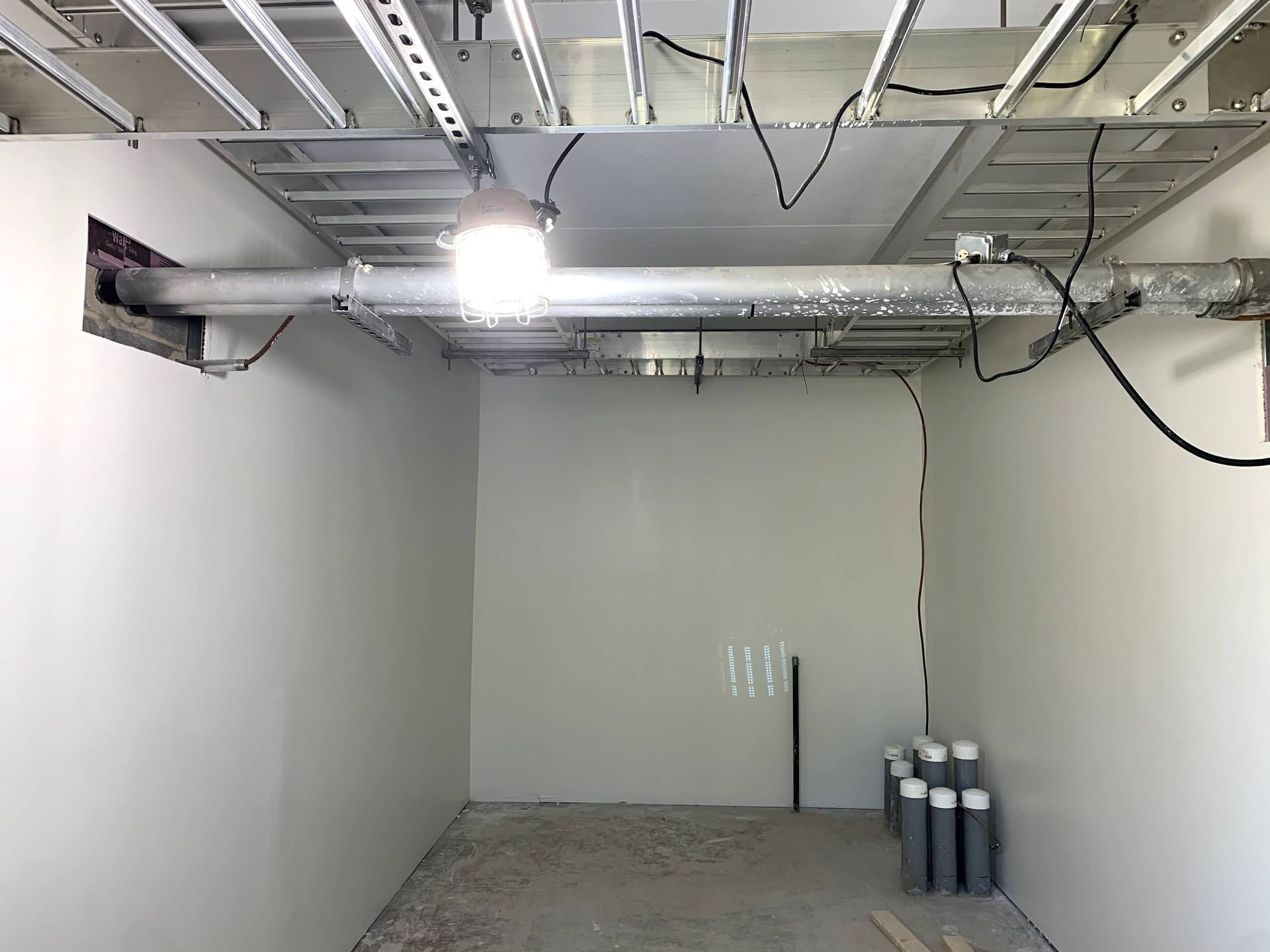 Empty, unfinished small room with white walls, exposed ceiling wiring and ductwork, and a single hanging light.