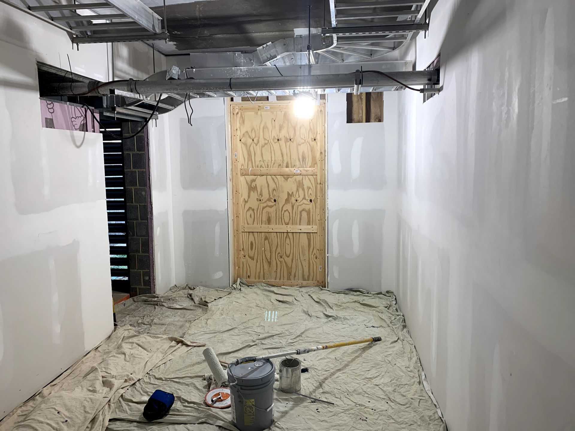 Interior room under construction with drywall, unfinished wooden door, and protective covering on the floor. Exposed ceiling beams and overhead light.