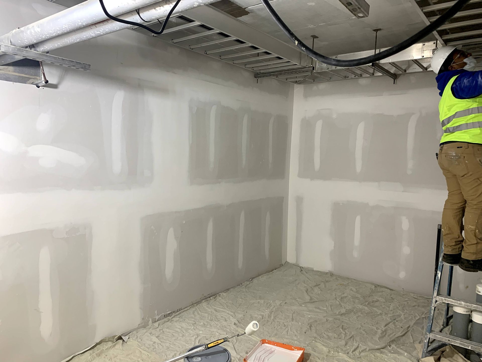 Construction worker on a ladder installing drywall in a room. Walls are white with gray drywall panels.