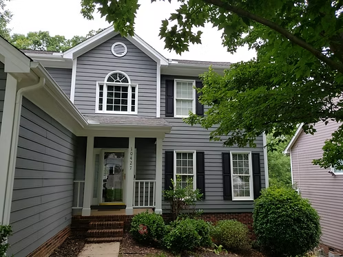 Gray two-story house with white trim, black shutters, and a small front porch, framed by trees and landscaping.