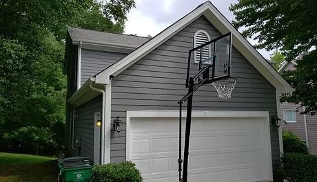 Gray house with white garage door, a basketball hoop, and green foliage.