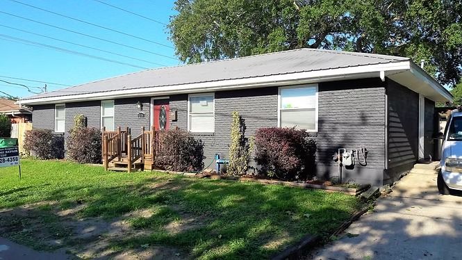 Black-painted house with a small porch and red door, bushes, and a grassy yard on a sunny day.